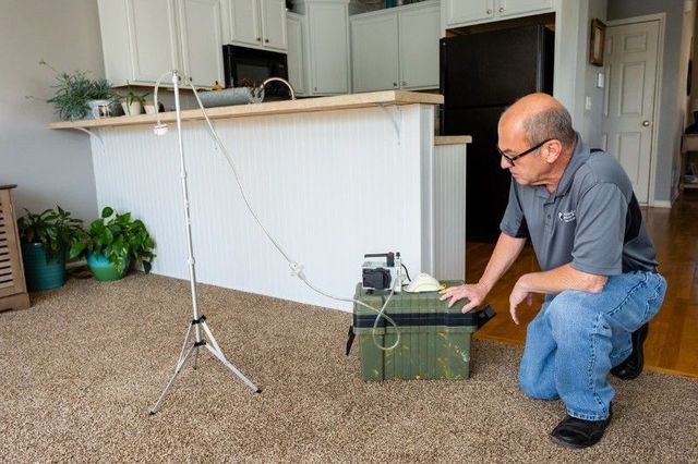 Man kneeling by a green equipment box in a carpeted room, adjusting a device on a tripod beside him.