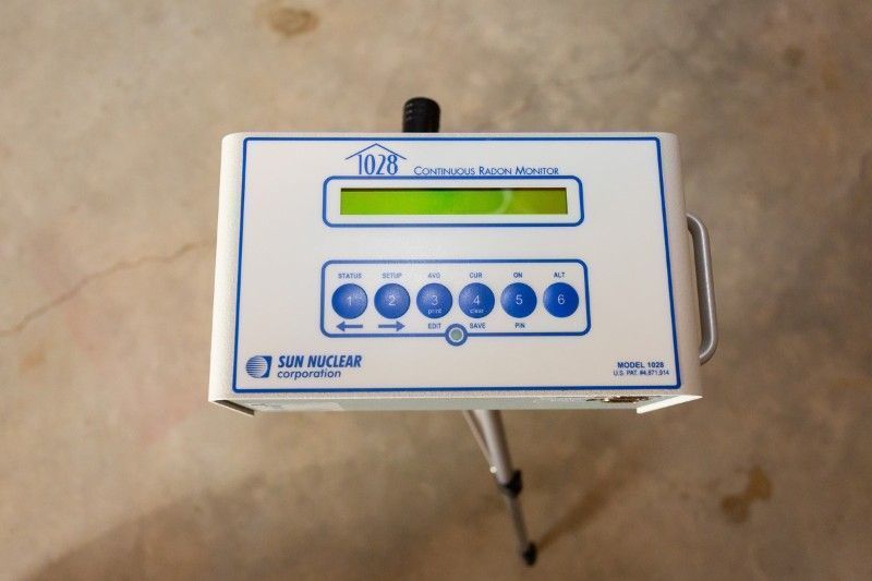 Digital control panel with a small screen and blue buttons on a stand, shown on a beige floor.