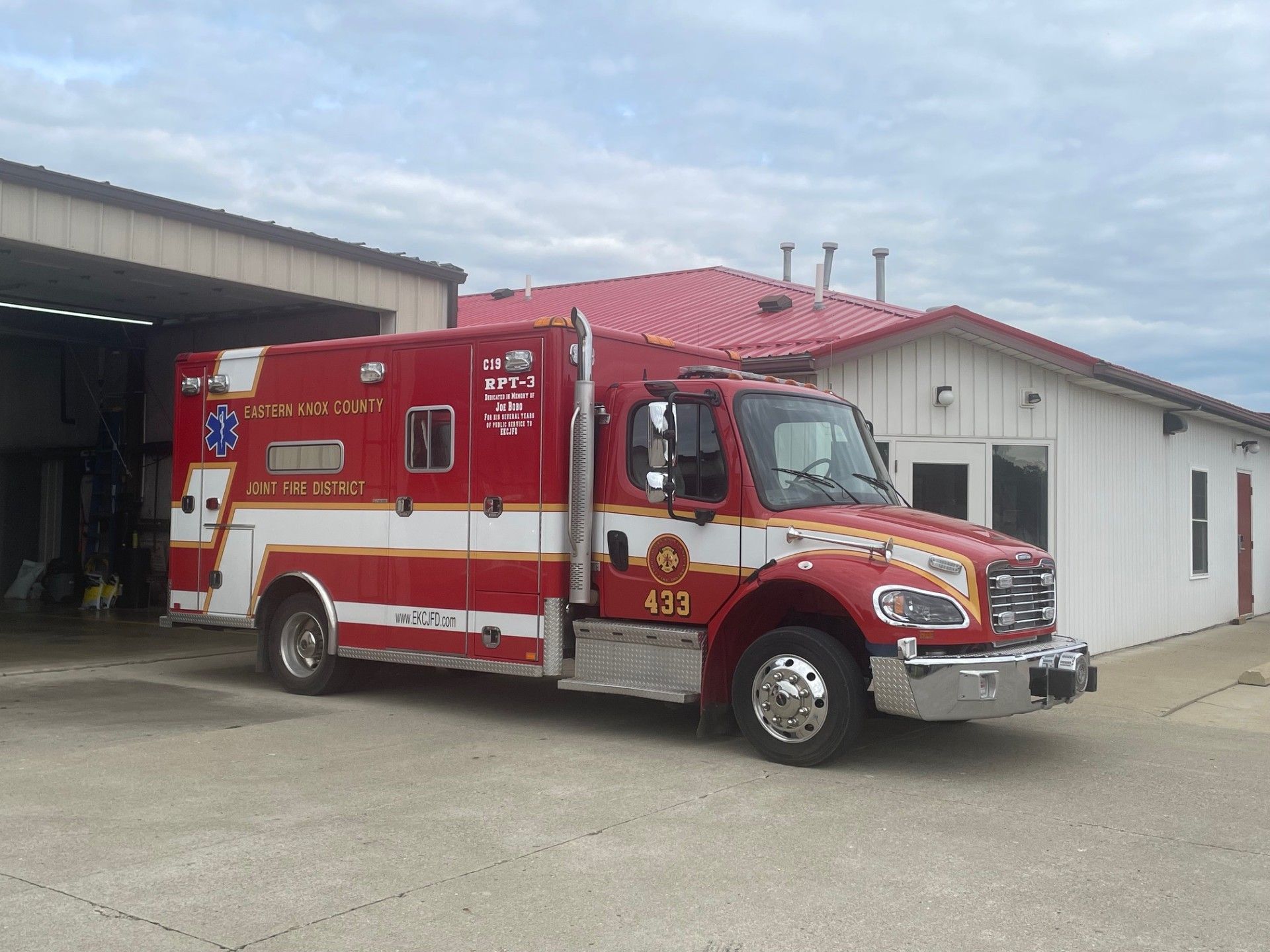 Red and white ambulance parked in front of a building with a red roof, displaying emergency service markings.