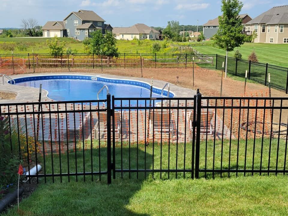 A backyard swimming pool with a stone deck surrounded by a black metal fence, overlooking a grassy residential area.