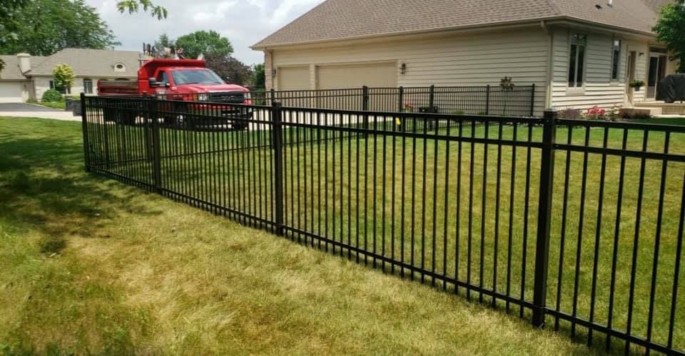 A black metal fence encloses a green grassy yard in front of a house, with a red dump truck parked in the driveway.