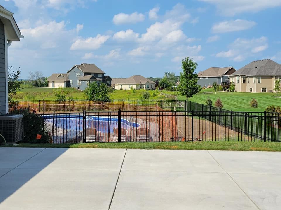 A backyard patio overlooking a swimming pool enclosed by a black metal fence, with suburban houses in the background.