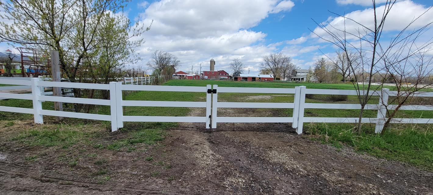 A white, three-rail wooden fence with a central double gate, set against a green field and a partly cloudy blue sky.