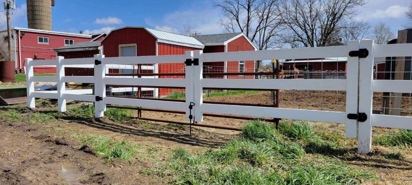 A white wooden three-rail fence stands in a grassy field in front of red farm buildings under a blue sky.