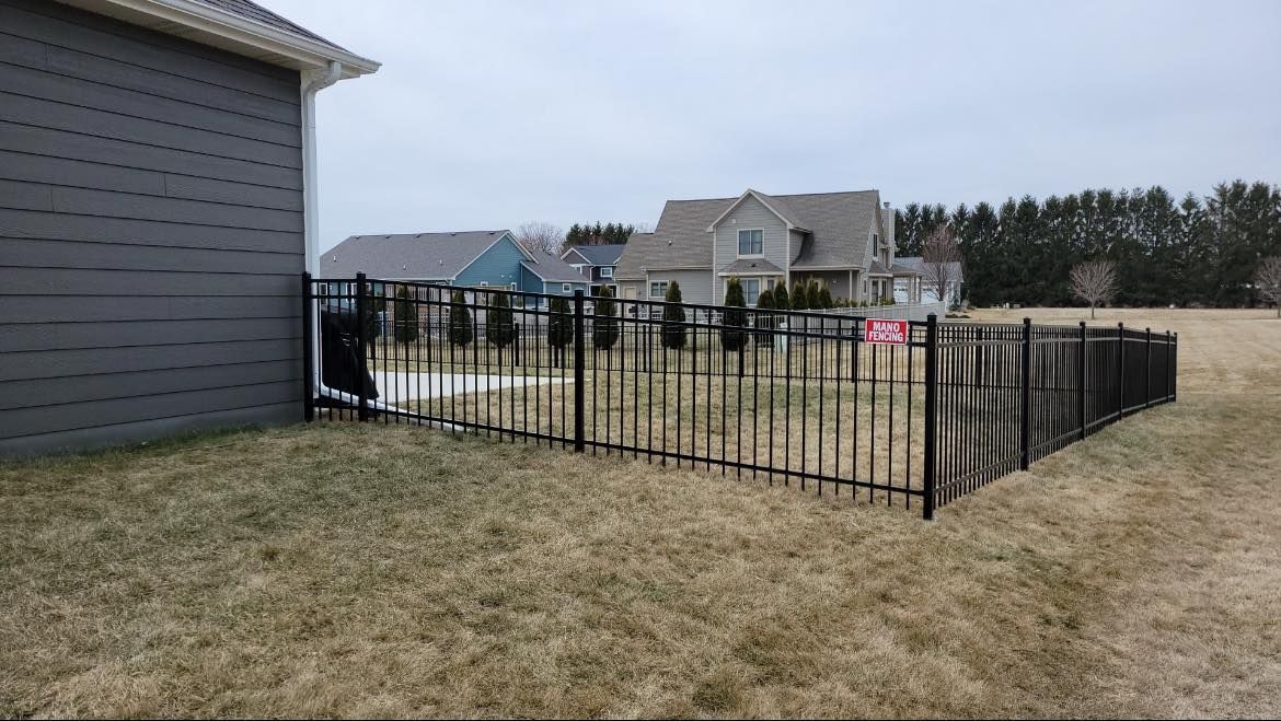 A backyard with a dark metal fence, dry grass, and a gray house exterior under a cloudy sky.