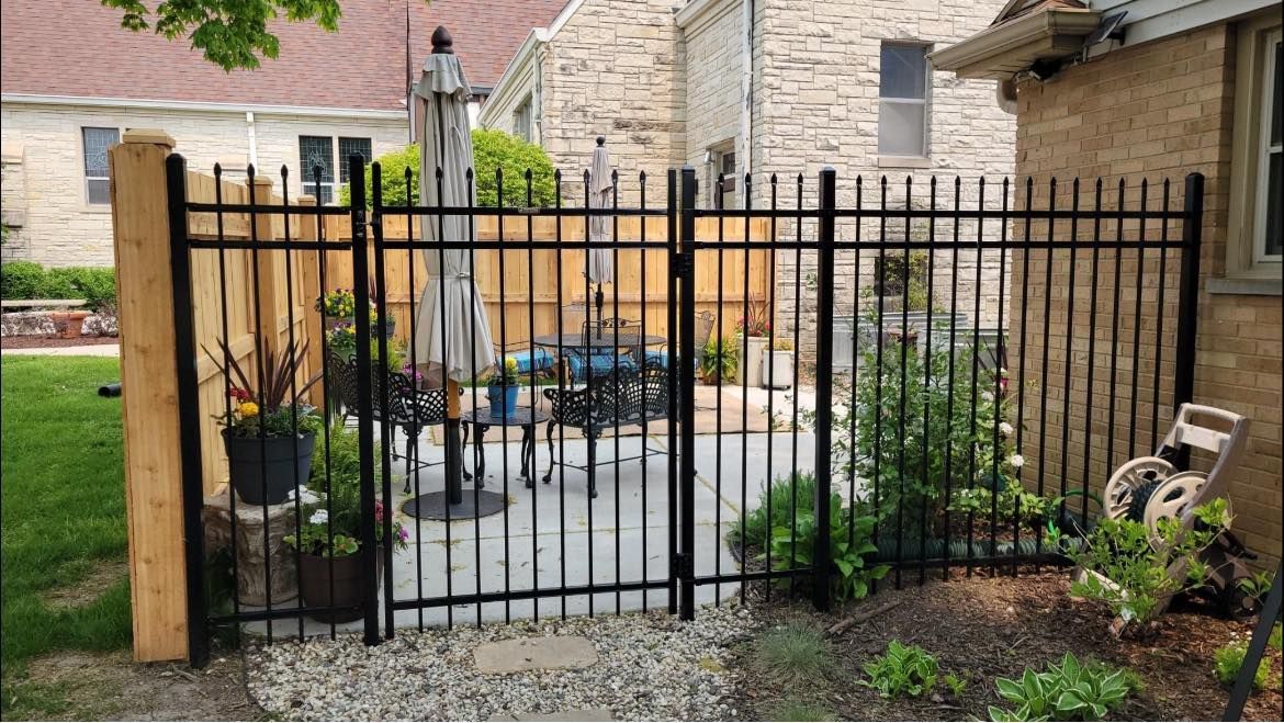 A black metal fence and gate surround a stone patio with outdoor furniture, potted plants, and a closed patio umbrella.