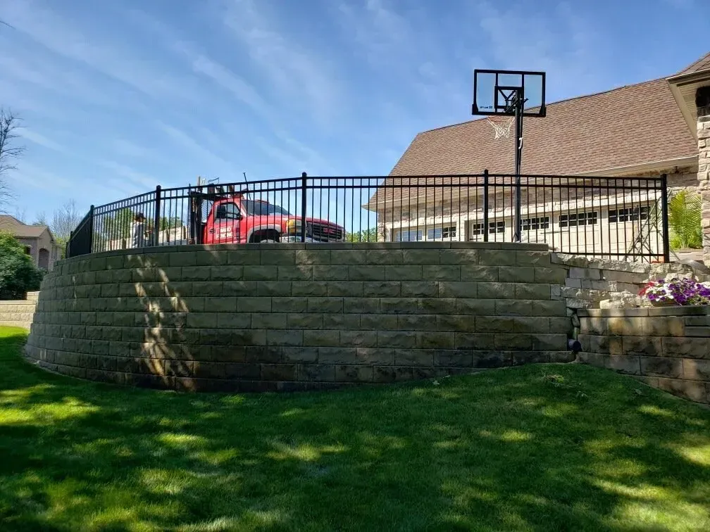 A red truck parked behind a black metal fence atop a curved stone retaining wall next to a house with a basketball hoop.