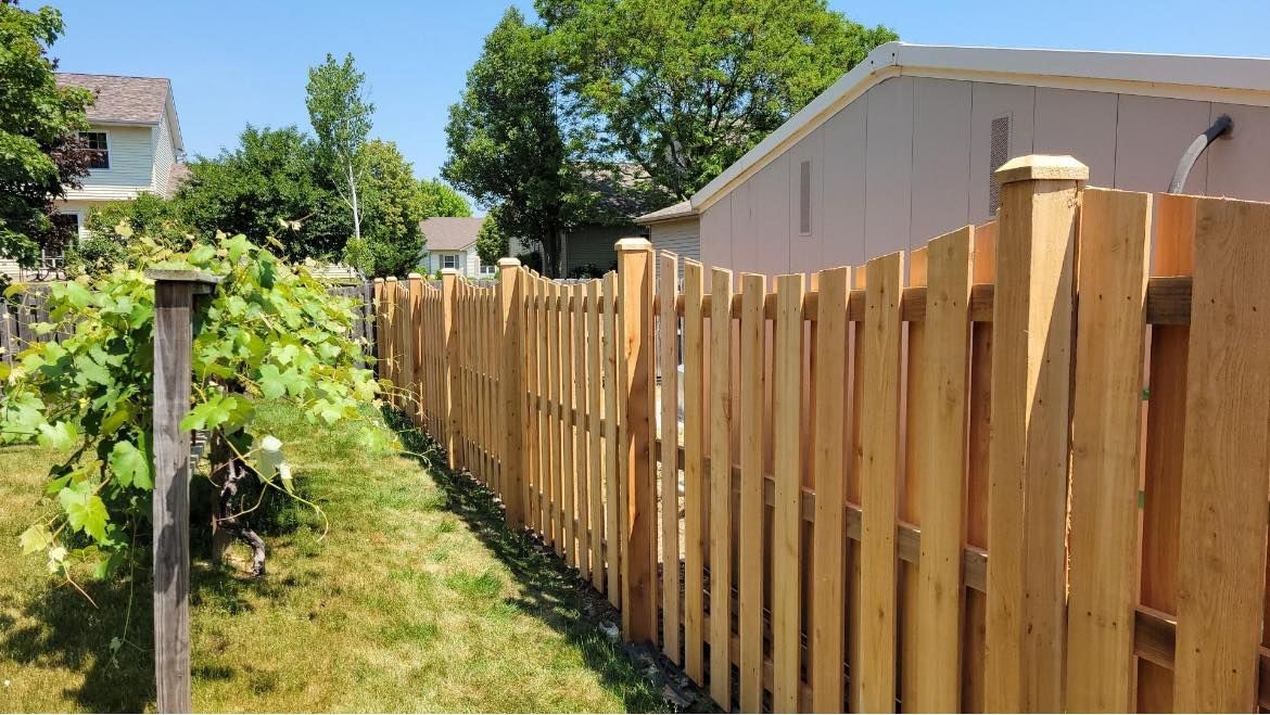 A new wooden fence stands along a grassy lawn next to green foliage on a sunny day.