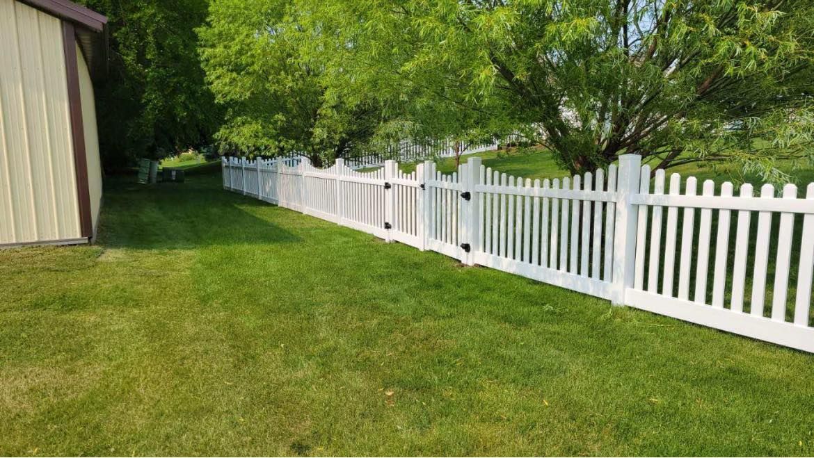 A white vinyl picket fence spans across a green lawn next to a light-colored building with trees in the background.