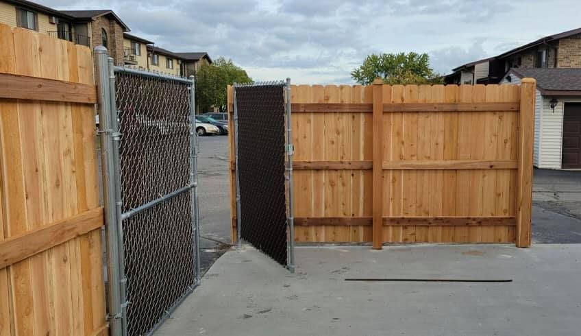 A chain-link gate stands partially open, nestled between wooden privacy fence sections in an outdoor parking area.