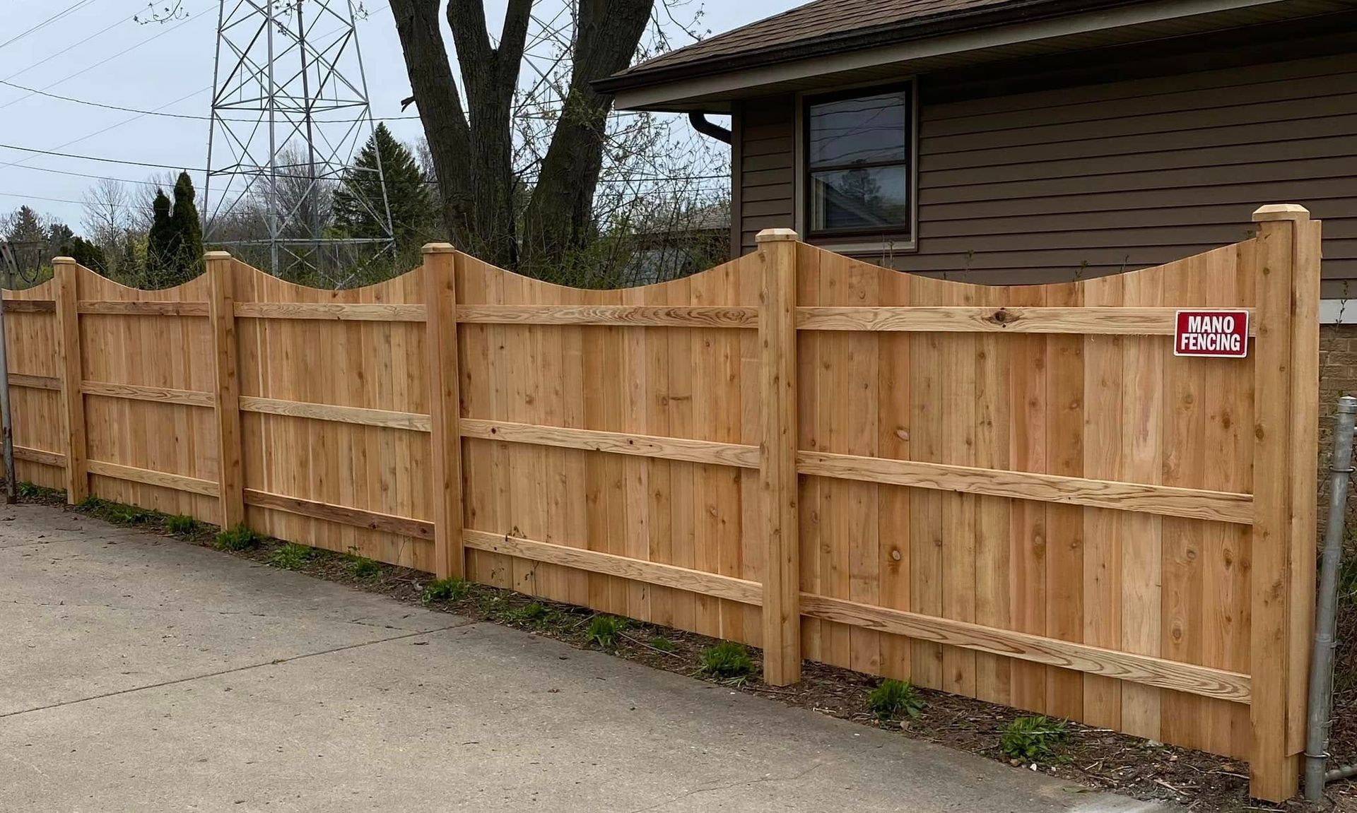A tall, scalloped-top wood privacy fence stands along a concrete driveway next to a house.