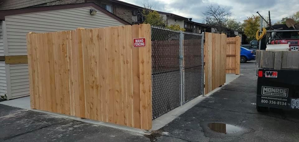 New cedar dumpster enclosure with a side gate and a chain-link fence panel, situated in a paved parking lot.