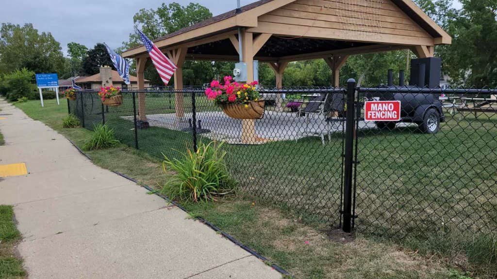 A wooden pavilion with a patio and a large grill trailer, situated behind a black chain-link fence with hanging flowers.
