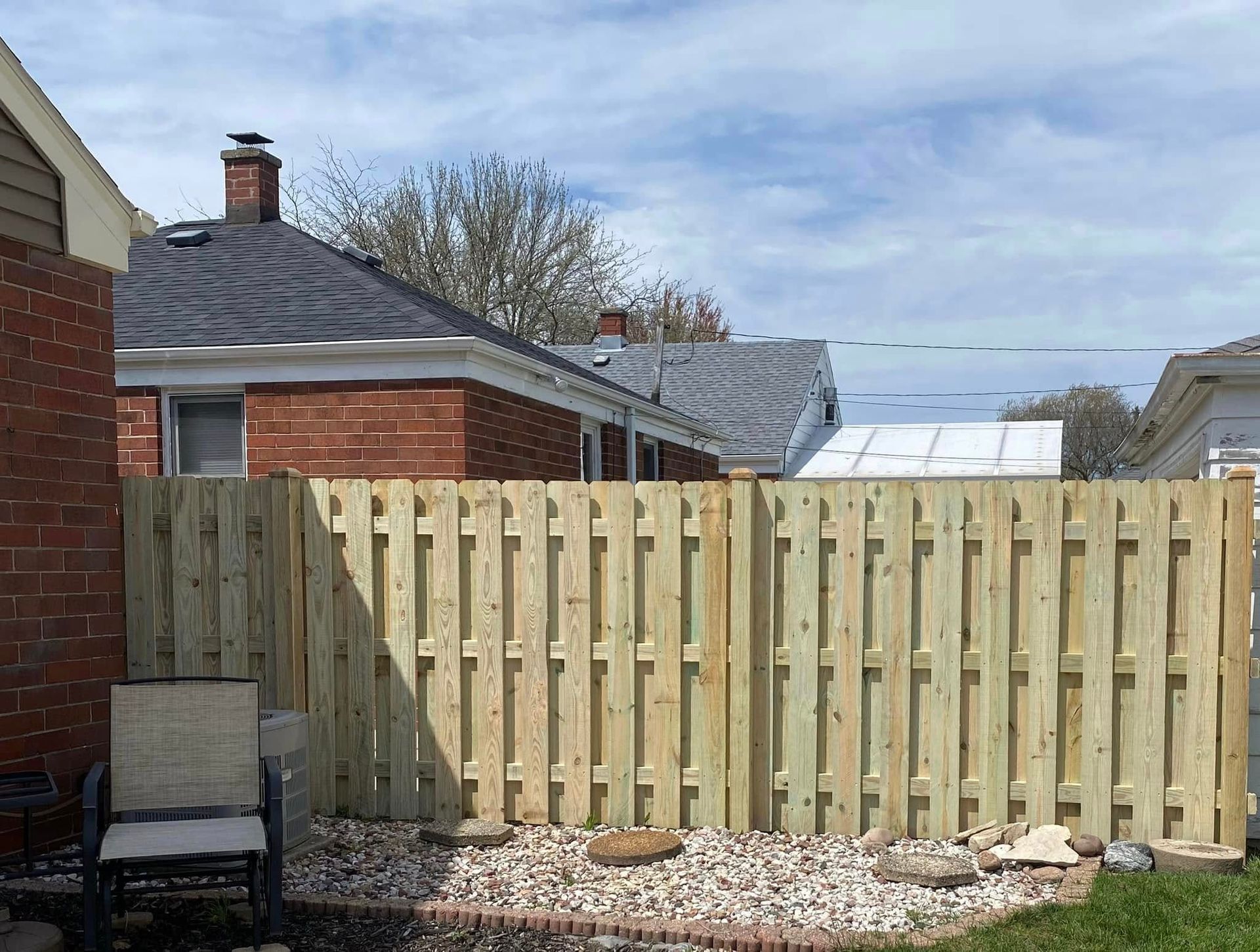 A newly installed light-wood privacy fence stands in a yard with a pebble-covered garden bed and a single chair.