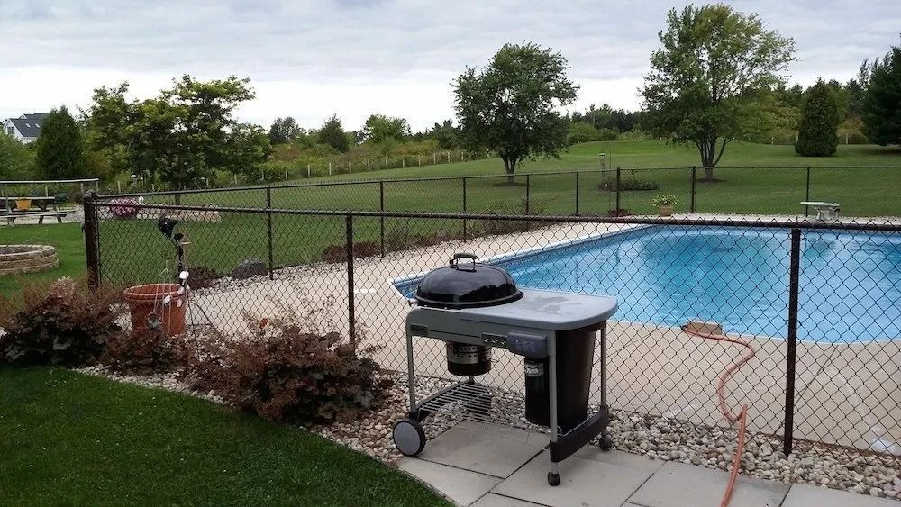 A portable charcoal grill sits on a concrete patio next to an outdoor swimming pool enclosed by a chain-link fence.