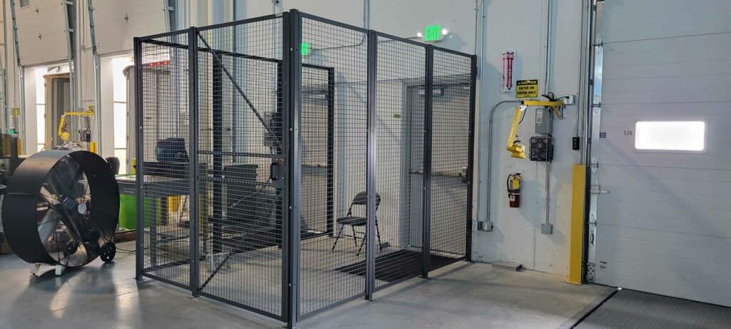A dark metal wire mesh enclosure stands in a warehouse near a large fan, a yellow support arm, and a building exit.
