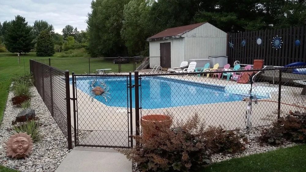 A backyard swimming pool enclosed by a chain-link fence, with a small shed and various lawn chairs in the background.