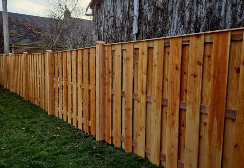 A light brown cedar wood fence with vertical pickets standing in a grassy yard on a cloudy day.