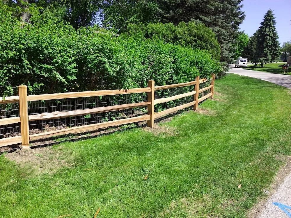 A wooden three-rail fence with wire mesh backing sits along a lush green hedge next to a lawn and paved path.
