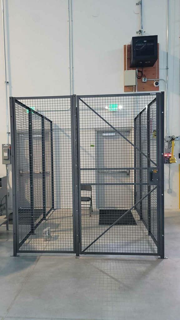 A black wire mesh security cage partition stands in an industrial warehouse setting in front of an emergency exit door.