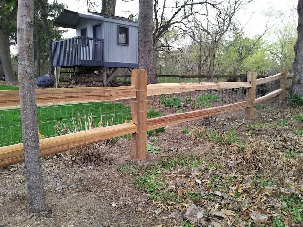 A wooden post-and-rail fence with wire mesh runs through a grassy yard toward a small elevated playhouse in the trees.