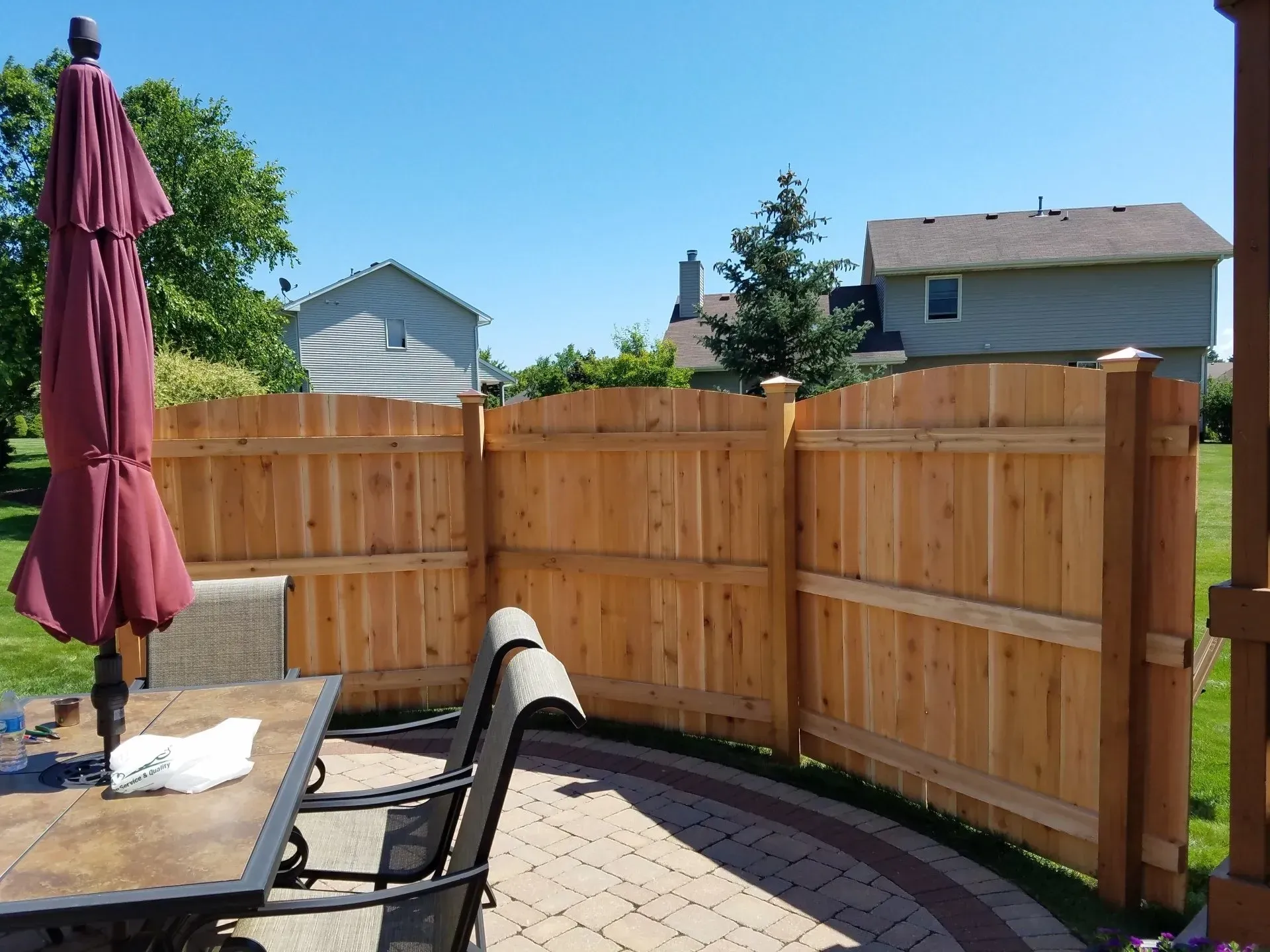 A wooden privacy fence surrounds a stone patio with a patio table, chairs, and a maroon umbrella on a sunny day.