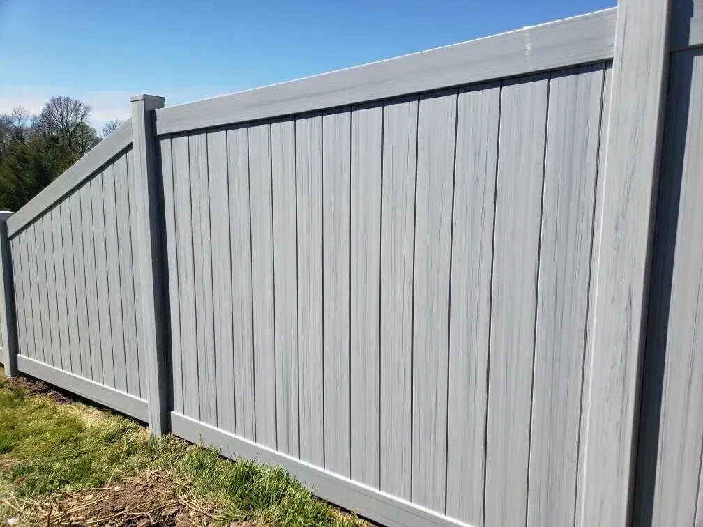 A grey vinyl privacy fence with vertical panels and a slanted transition section against a bright blue sky.