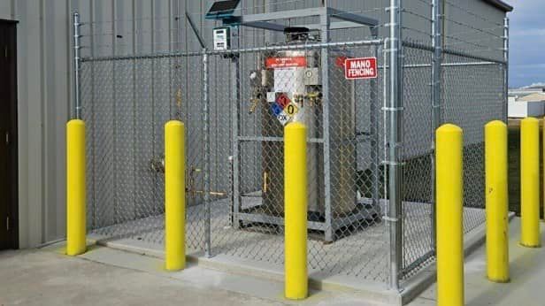 A metal storage tank enclosed in a chain-link fence, protected by yellow safety bollards outside a building.