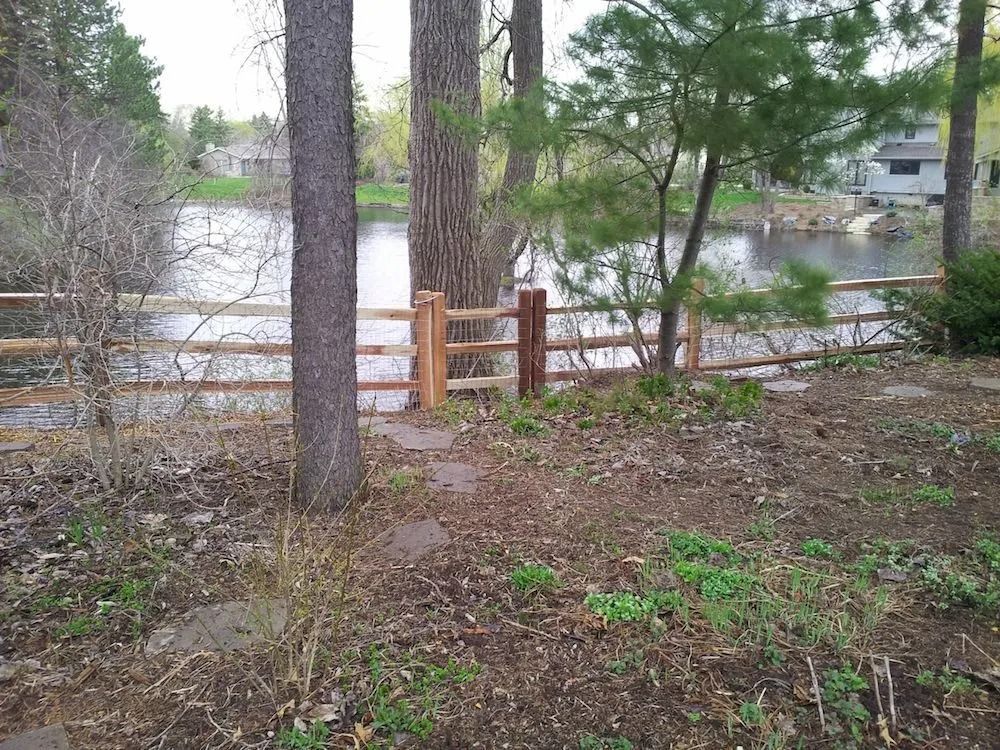 A wooden split-rail fence runs along the edge of a pond, surrounded by trees and a dirt yard in early spring.