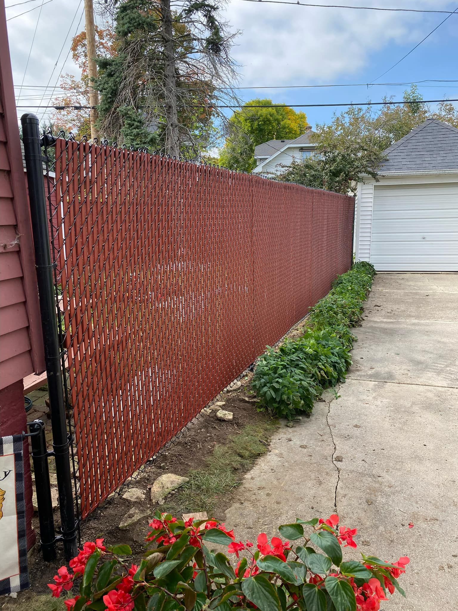 A tall red chain-link fence with privacy slats runs alongside a concrete driveway near green shrubs and a garage.