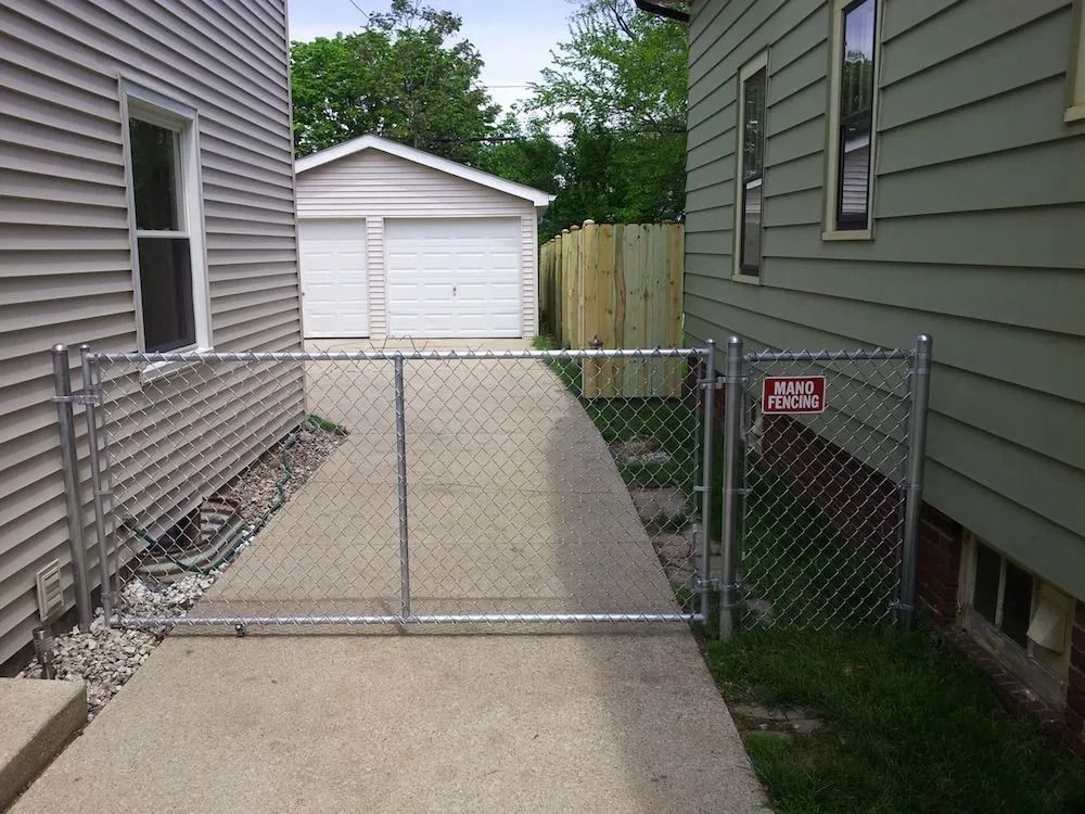 A metal chain-link gate stretches across a concrete driveway between two residential houses with a garage in the back.
