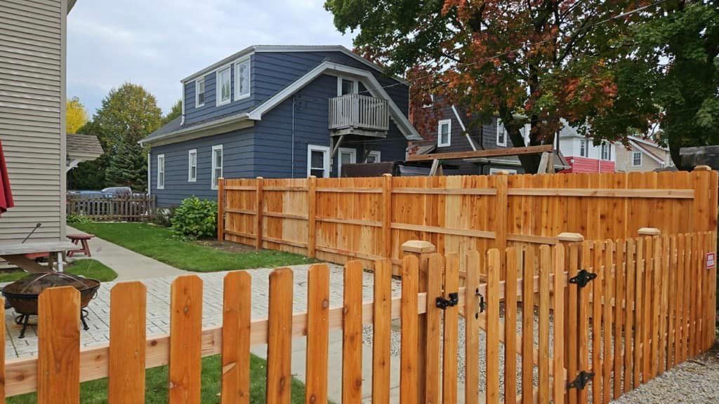 A new wooden picket fence encloses a patio area with a paved walkway, in front of a blue two-story house.