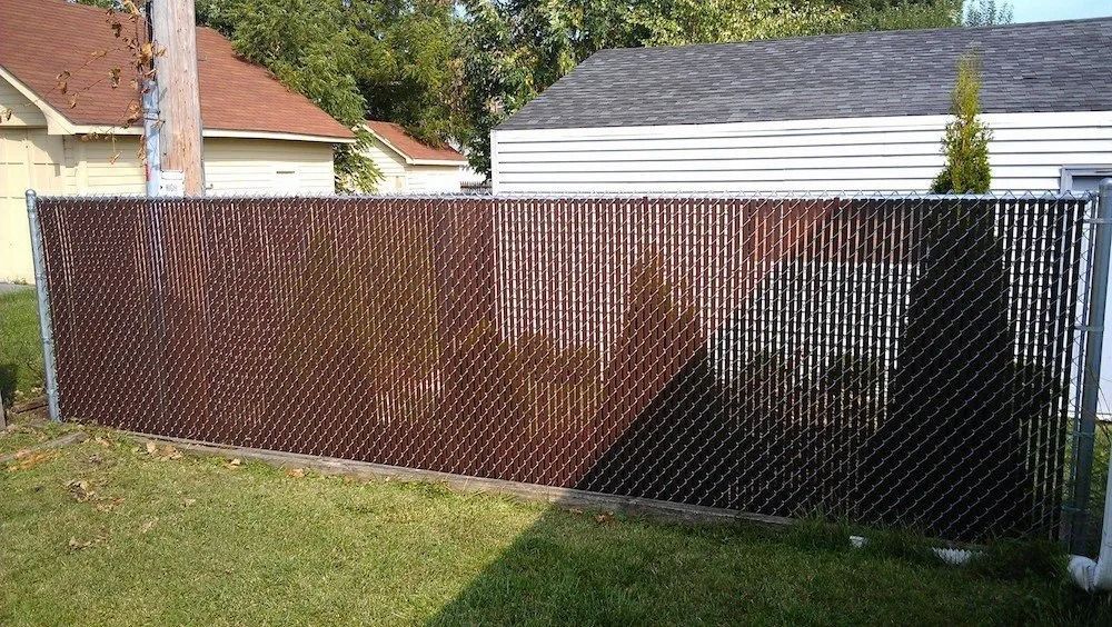 Brown plastic privacy slats woven into a chain-link fence in front of houses and a small tree.