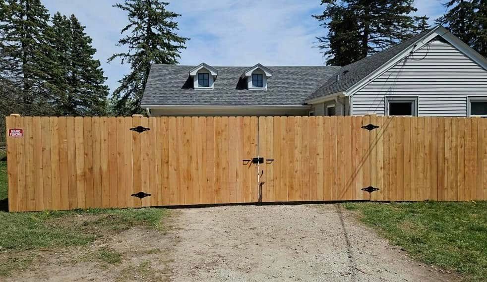 A large wooden double gate sits across a gravel driveway in front of a white house with a grey roof.