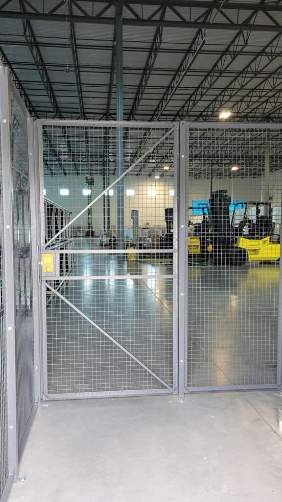 A grey metal safety gate with wire mesh panels stands in a warehouse with a forklift visible in the background.