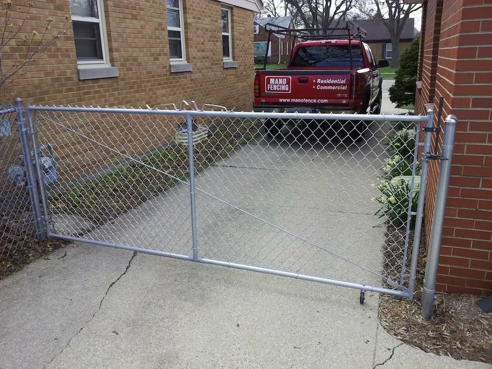 A chain-link gate blocks a residential driveway, with a red truck parked behind it next to a brick house.