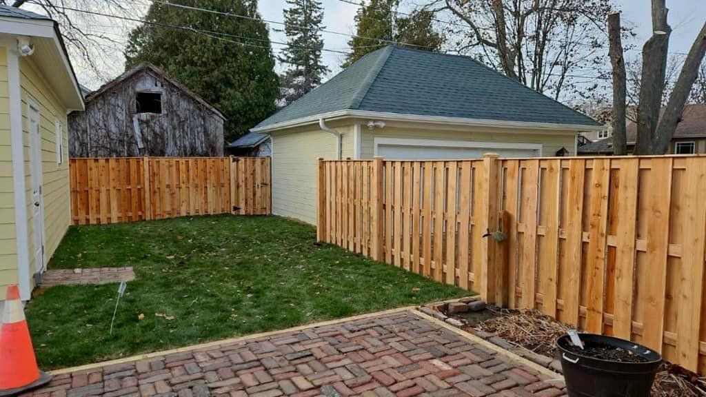 A backyard with a red brick patio, green grass, new cedar fence, and a yellow garage under an overcast sky.
