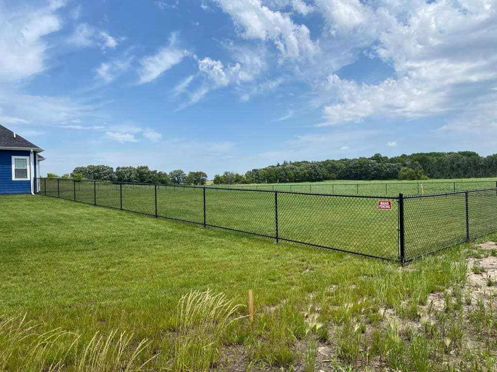 A blue house with a black chain-link fence bordering a large, green grassy field under a sunny blue sky.