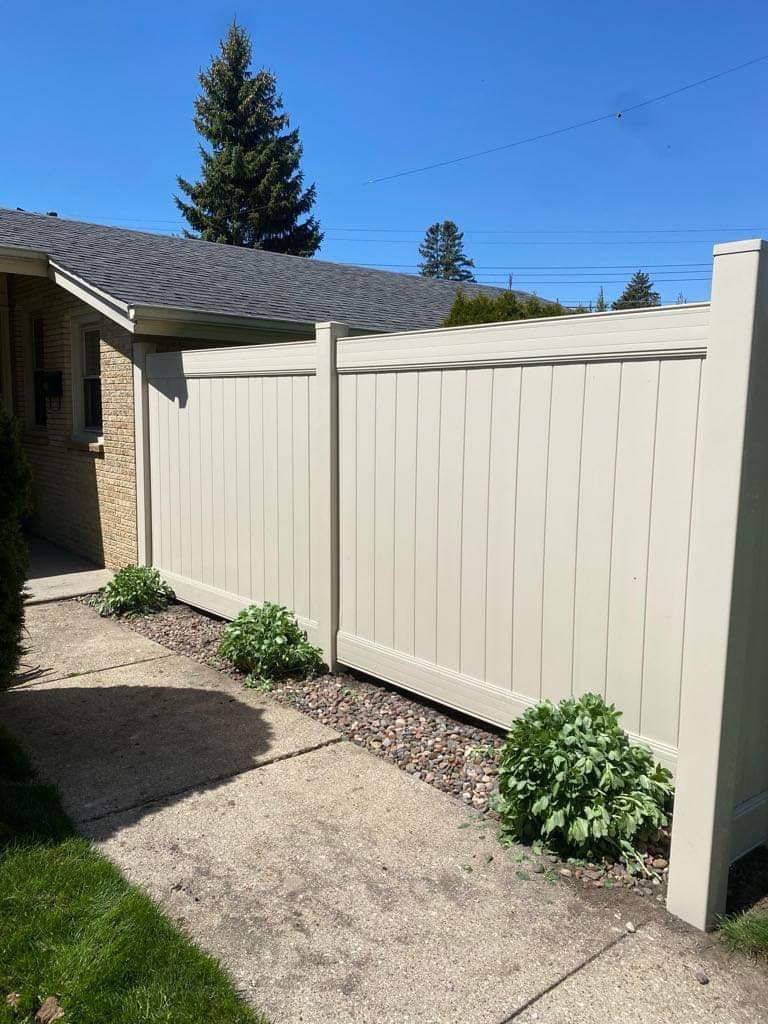 A tan vinyl privacy fence runs alongside a concrete walkway next to a brick building under a clear blue sky.
