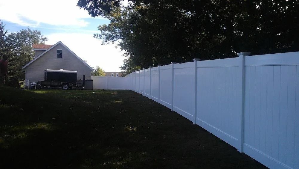 A long, white vinyl privacy fence lines the edge of a grassy backyard, with a house and trailer visible in the distance.