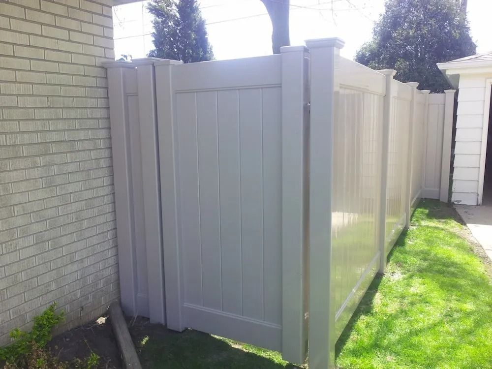 A tan, solid-panel vinyl privacy fence stands outdoors next to a light-colored brick house and a grassy lawn.
