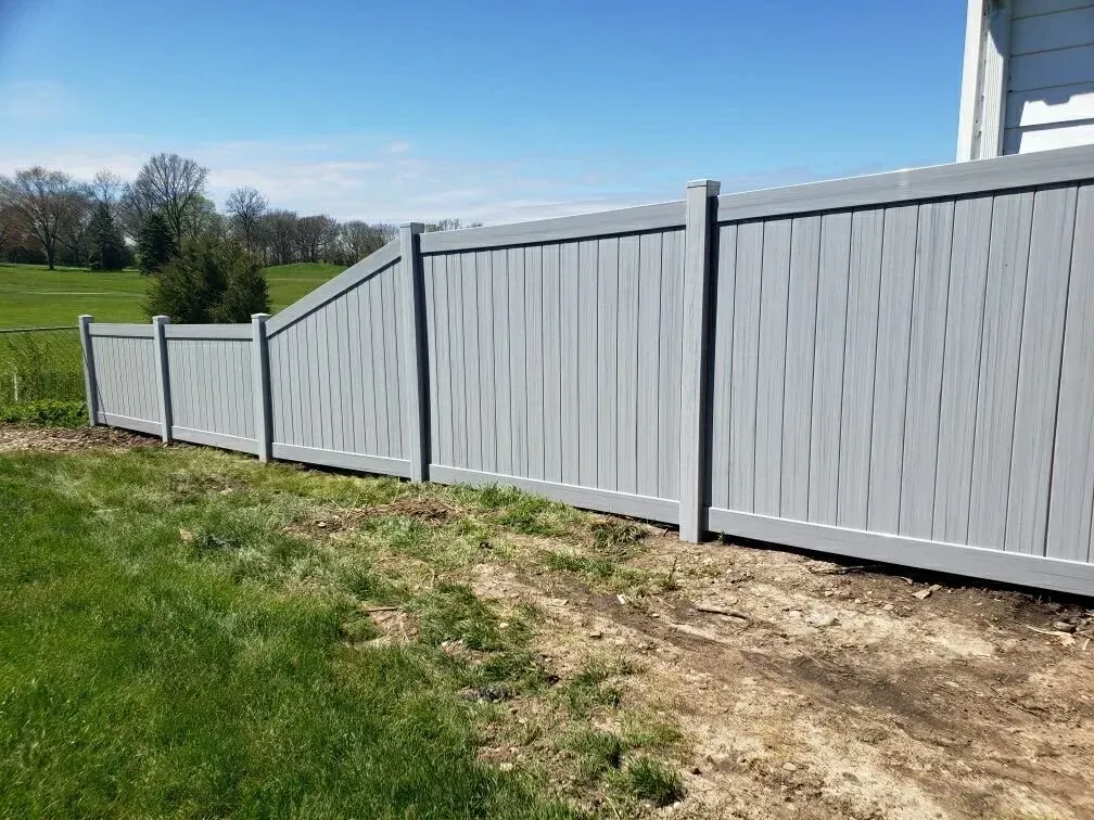 A grey vinyl privacy fence steps down in height along a grassy slope next to the side of a house.