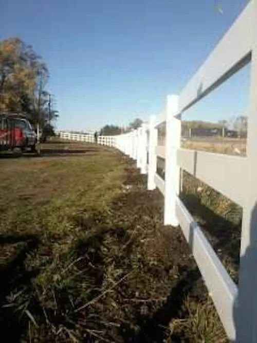 A long, white, three-rail vinyl fence curves across a grassy field under a clear blue sky.