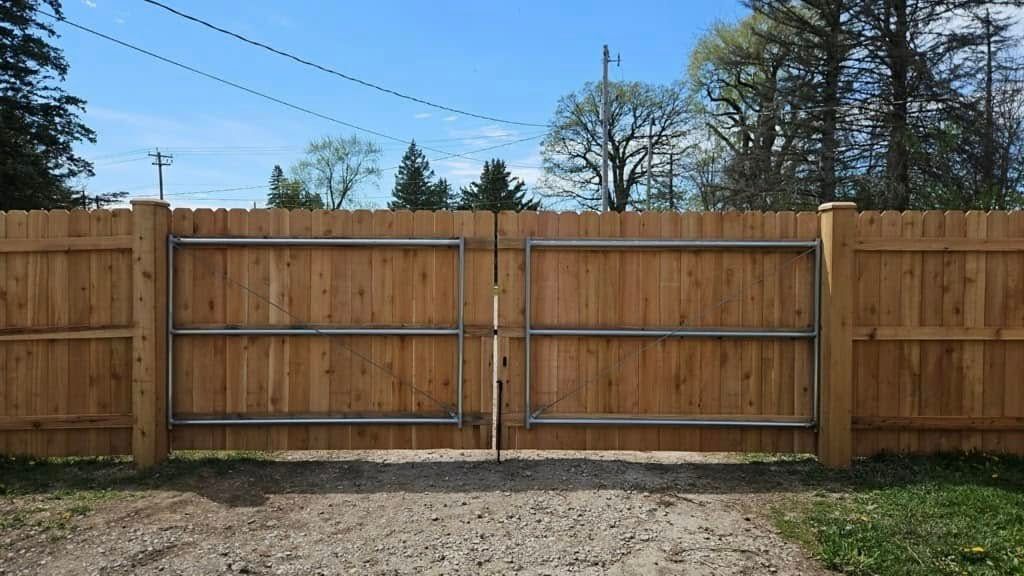 Double wooden driveway gate with metal frames set in a wooden privacy fence outdoors on a gravel driveway.