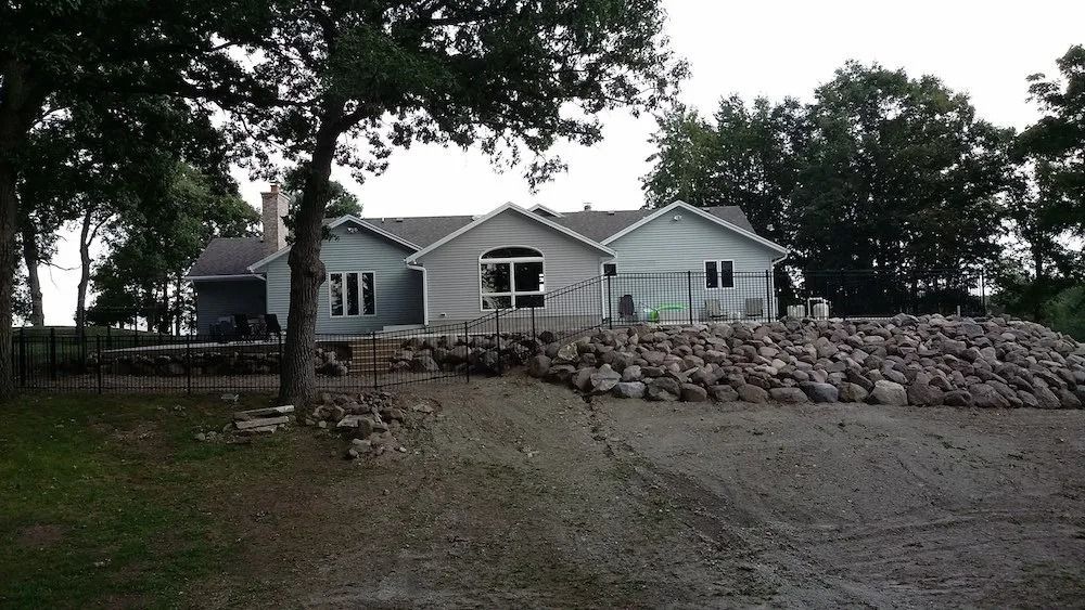 Light gray single-story house with a rock retaining wall and gravel yard, surrounded by trees.