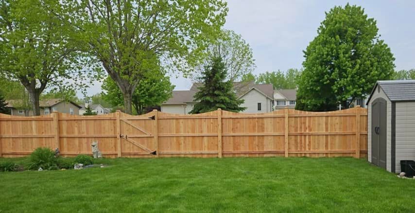 A wooden privacy fence with an arched gate runs across a green backyard lawn, flanked by trees and a shed.