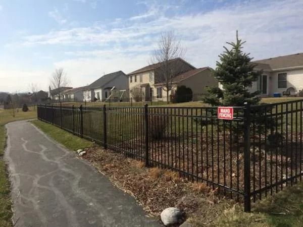 A paved sidewalk runs alongside a black metal fence with a no parking sign, bordering a line of suburban houses.
