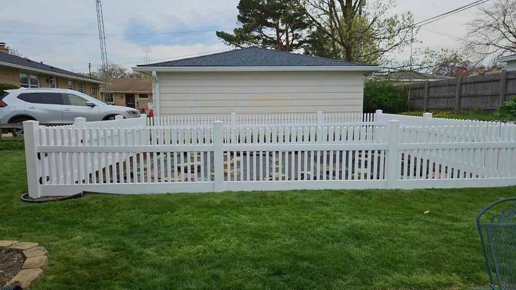 A white picket fence encloses a backyard garden area in front of a detached garage on a suburban property.