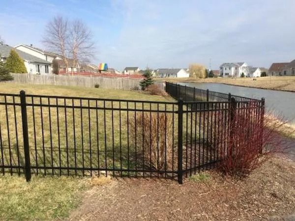 A black metal fence encloses a grassy backyard next to a small pond in a suburban neighborhood.