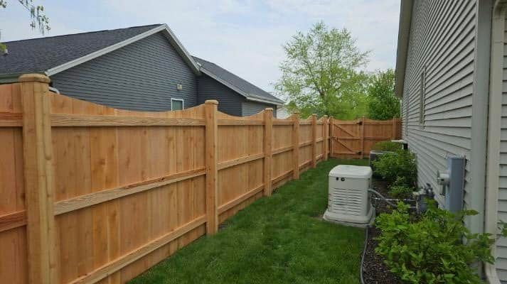 A new wooden privacy fence with a scalloped top border runs along a lawn beside the side wall of a residential house.